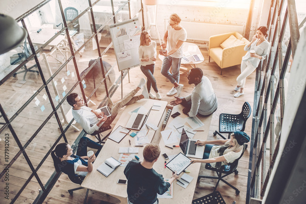 A conference room full of diverse young workers having a productive meeting. They have laptops on their laps and the table has assorted papers, demonstrating that they're productive.