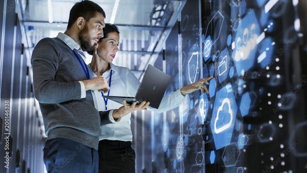 An IT professional with a laptop standing in a data center with another employee. They are pointing at the racks of servers, where graphical overlays show icons of a shield, folders, a globe, and other various iconography related to servers and cybersecurity.