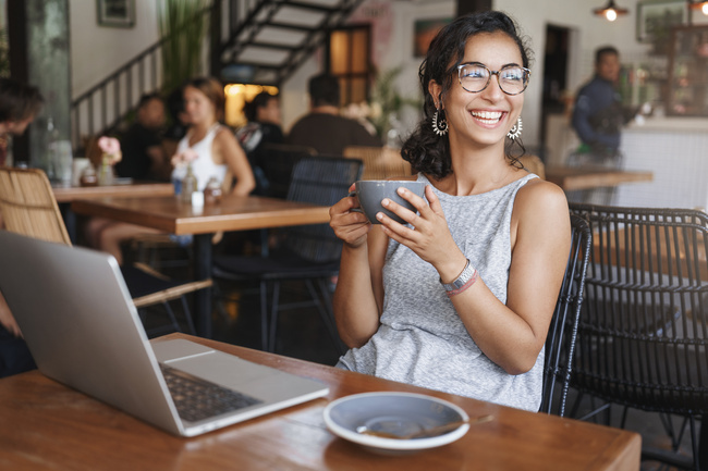 A Woman sitting in a busy coffee shop with a cup of coffee in her hand. She's looking to the side and smiling, and her laptop is on the table in front of her.
